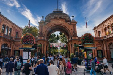 Tivoli Bahçeleri 'ne giriş, Kopenhag, süslenmiş bir kemer yolu üzerinde TIVOLI, kırmızı tuğla ve bir saat. Çeşitli kıyafetlerle dolu canlı bir kalabalık, sallanan çiçekler ve parçalı bulutlu bir gökyüzünün altında bayraklar..