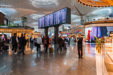 İstanbul Havalimanı 'nda, gezginler ve BVLGARI gibi lüks mağazalarla süslü bir terminal mırıldanıyor. Bir muhafız, etrafı sanat ve kapı işaretleriyle çevrili bir uçuş panosuyla tarıyor..