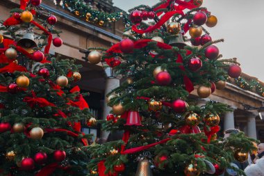 Şenlikli Londra sahnesi, yumuşak ışıklandırma altında kırmızı ve altın takılar, kurdeleler ve büyük süslemelerle süslenmiş zengin Noel ağaçları. Bina cephesinde çelenkler ve altın ışıklar var..