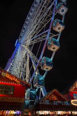 Karla kaplı Noel tezgahlarının sıcak ışıklarına karşı serin maviyle aydınlatılmış, yüksek bir dönme dolabı olan şenlikli bir Londra sahnesi..