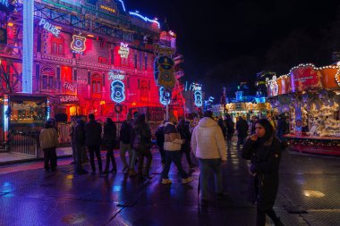 Renkli, yeni ışıklandırılmış polis karakolu temalı canlı bir Londra Noel piyasa sahnesi. İnsanlar, ıslak zeminde kırmızı ve mavi ışıkların karışımı altında bir atlıkarıncanın yanındaki şenlikli atmosferden hoşlanırlar..
