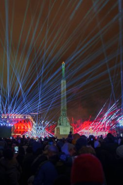 Rigas Özgürlük Anıtı 'nda canlı bir gece sahnesi. Renkli ışık huzmeleriyle yeşille aydınlatılmış, kışın giyinmiş bir kalabalık Letonya' nın Bağımsızlık Günü 'nü kutluyor..