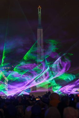 Riga, Letonya 'da, Bağımsızlık Günü kutlamaları sırasında Özgürlük Anıtı' ndaki yeşil ve mor lazer gösterisini izleyen bir kalabalığın olduğu canlı bir gece sahnesi..