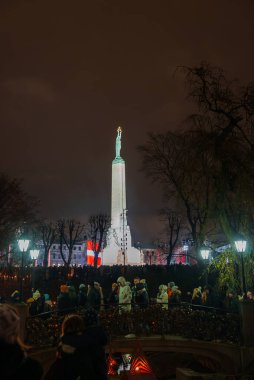 Letonya 'daki Rigas Özgürlük Anıtı' nda, aydınlanmış bağımsızlık sembolünün etrafında toplanan siluetli kalabalıklarla dolu bir festival gecesi sahnesi.