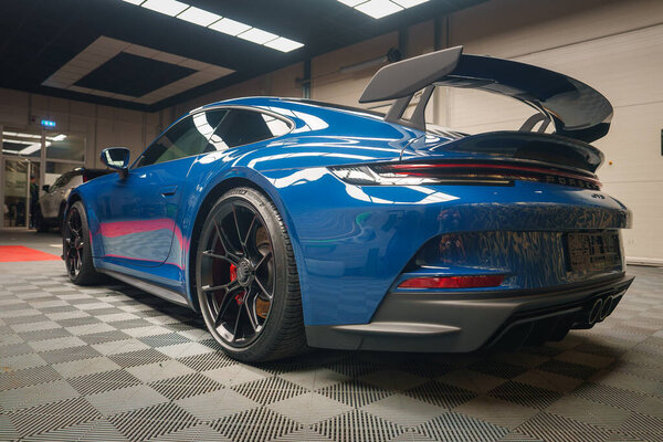 A gleaming blue Porsche 911 GT3 with a prominent rear wing and Porsche lettering is showcased indoors, reflecting lights on its polished surface and sporty alloy wheels with red calipers.