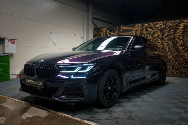 Parlak LED farları ve böbrek ızgarası olan parlak siyah bir BMW garaja park edilmiş, cilalı dış ve siyah jantlarından yansıyan çevre ışığını duvar tipi bir duvara yansıtıyor..