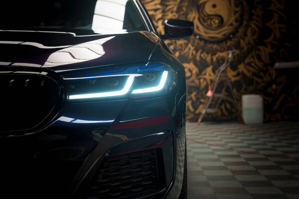 Closeup of a polished black Audis front end with LED headlights on, reflecting ambient light against a patterned floor and artistic mural backdrop, likely in a luxury car tuning garage.
