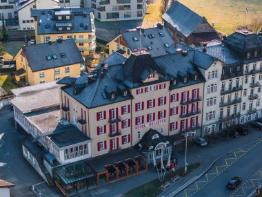 Aerial view of the elegant Hotel Bellevue in Engelberg, showcasing its white facade with red shutters, surrounded by a serene town with diverse architecture and a tranquil atmosphere. clipart