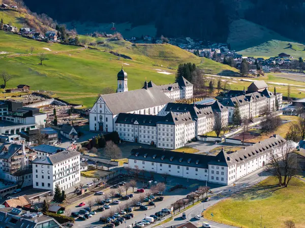 Luxurious hotel complex nestled in Engelberg valley, featuring white facades, dark roofs, and a historic church with a clock tower. Surrounded by alpine scenery, popular with visitors.
