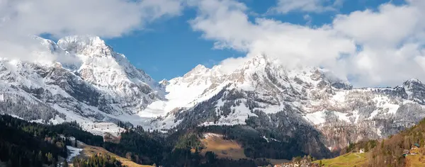 スイスのエンゲルベルクのパノラマビューは 雪に覆われた山々 常緑の木々 部分的に曇った空の下にアルペンビルを持つ谷を示し 自然の壮大さを強調しています — ストック写真