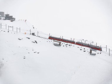 Aerial view of a vibrant red train navigating through the snowy landscape of Zermatt, Switzerland, on an elevated track amidst ski tracks and a nearby ski resort structure, under whiteout conditions. clipart