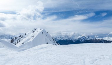 Hava görüntülerinde Verbier, Switzerlands karlı manzarası, kayak pistleri, tepe noktası ve bulutlu bir gökyüzünün altındaki Alpler görülüyor. Kış cazibesi el değmemiş kar ve yumuşak bulut gölgelerinde.