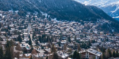 Aerial view of Verbier, Switzerland, showcasing snow covered chalets and buildings typical of alpine architecture, nestled in a forested mountain landscape with the Swiss Alps in the background. clipart