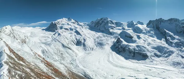 İsviçre 'deki Zermatt kayak merkezinin çarpıcı manzarası açık mavi gökyüzünün altında engebeli, karlı zirveleri gösteriyor. Güneş ışığı dağları özetliyor, dingin, el değmemiş dağların güzelliğini ortaya çıkarıyor..