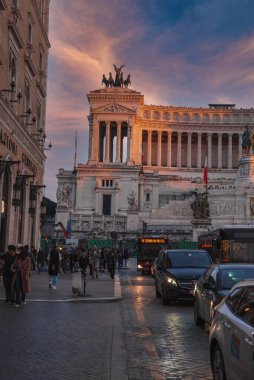Roma kenti alacakaranlıkta Altare della Patria anıtı, kaldırım taşı caddesinde yayalar ve yol kenarında Assicurazioni Generali de dahil olmak üzere ticari binalar.