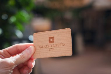 Close up of hand holding wooden card with PALAZZO RIPETTA ROMA engraved, hinting key or ID for luxury hotel in Rome. Blurry green garden background. Bright natural light hints daytime. clipart