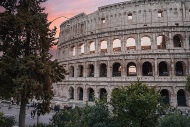 Roma, İtalya 'daki antik amfitiyatroda, ikonik Colosseum dış görünüşü, kemerler, taş duvarlar ve yeşillikler sergileniyor. Turistler ve yerliler alacakaranlıkta tarihi mekanın yakınında yürürken, dingin bir hava yakaladılar..
