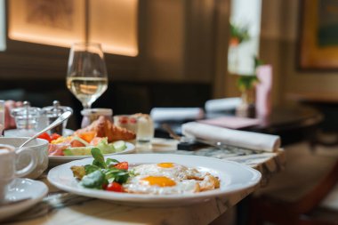 Roma 'daki lüks bir otelde lüks kahvaltı. Yumurtalar, domatesler, beyaz tabakta yapraklı yeşiller. Arka planda bir bardak beyaz şarap. Klasik zarif tasarım..