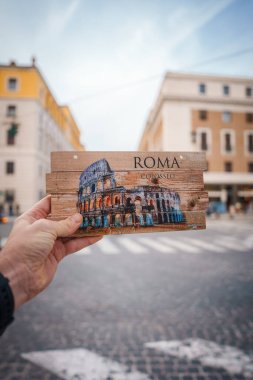 Üzerinde Colosseum sanat eserleri, ROMA ve COLOSSEO olan ahşap bir kartpostal taşıyordu. Klasik İtalya hatırası. Bulanık şehir arka planı ve tarihi binalar.