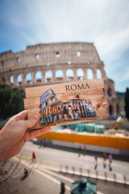 Roma 'daki Colosseum kartpostalı gerçek Colosseum ile aynı hizada. ROMA ve COLOSSEO kartpostalda. Arka planda gerçek bir dönüm noktası, güneşli bir sokak manzarası yayaların altında..