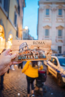 Kolezyum resimli ahşap kartpostalla el ele tutuşmak. ROMA üstte, COLOSSEO altta. Roma 'da kentsel unsurlarla arka plan sokak sahnesi.