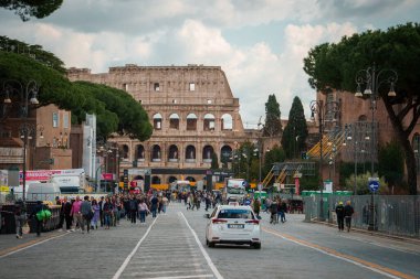 Arka planda Colosseum 'un olduğu hareketli sokak sahnesi, muhtemelen Roma, İtalya' da. Kalabalık kaldırımlar, beyaz araba sürüş, bulutlu gökyüzü, yoğun tarihi alan.