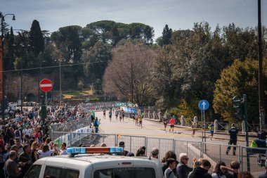 Road race event on a sunny day in an urban setting, possibly a city park. Spectators, runners, police vehicle suggest Rome, Italy. Large crowd, organized route with blue lines, lively atmosphere. clipart