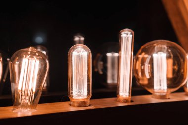 Collection of various shaped illuminated light bulbs emitting warm, amber glow on wooden surface. Filaments resemble squirrel cage type. Dark background creates cozy atmosphere. Location Unknown. clipart