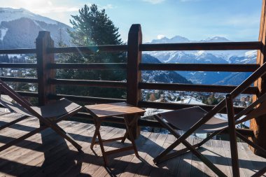 Tahta balkondan sakin dağ manzaralı sandalyeler ve dağlara bakan bir masa. Parlak güneş, karla kaplı dağlar, yemyeşil ağaçlar. Avrupa alp bölgesi.