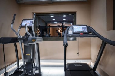 Solunda eliptik, sağında koşu bandı olan iki TechnoGym kardiyo makinesi olan modern bir spor salonu. İyi aydınlatılmış odanın yansıması camdan görülüyor..