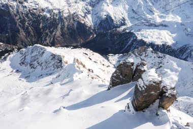 İsviçre 'deki Murren kayak merkezinin nefes kesici manzarası. Kar yamaçları, engebeli araziler, ön planda kaya oluşumları, dik dağlar, açık mavi gökyüzü. Kış sporları için ideal.