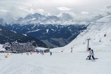Kış sporları aktiviteleriyle karlı dağ manzarası. Alp köyündeki snowboardcular, kayakçılar, chaletler. Majestic Peaks, kayak kaldırma sistemi, eğlence sahnesi. Popüler bir kayak merkezinde.
