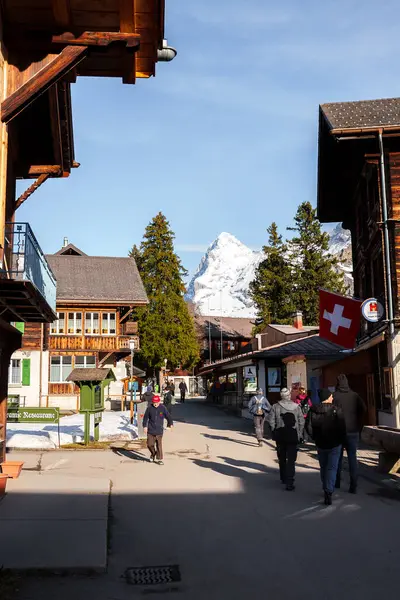 İsviçre, Murren 'de geleneksel dağ evi tarzı binalar, İsviçre bayrağı, caddesi ve yürüyen insanlar, görkemli kar örtülü dağlara karşı dizilmiş..