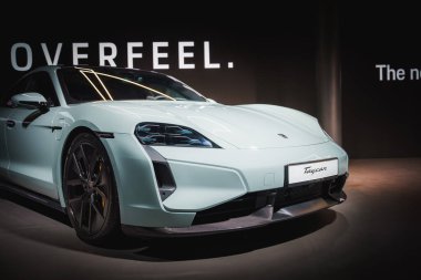 Beyaz Porsche Taycan elektrikli spor arabası, karanlık bir arkaplan önünde OVERFEEL ve yenisi kapalı alandaki profesyonel bir etkinlikte sergilendi.