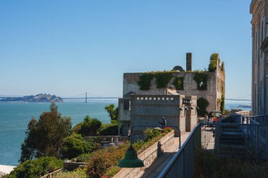 San Francisco, ABD 'deki cezaevi kompleksinde aşırı gelişmiş bitki örtüsüyle tarihi Alcatraz Adası manzarası. Ziyaretçiler körfez köprüsü ve mavi gökyüzü arka planıyla bölgeyi keşfediyorlar.