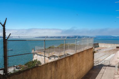 San Francisco, ABD 'deki Alcatraz hapishane manzarası. Resim dışarıyı gösteriyor. Zincirli tel örgüler ve körfez manzaralı. Arka planda sisle kaplı Golden Gate Köprüsü.