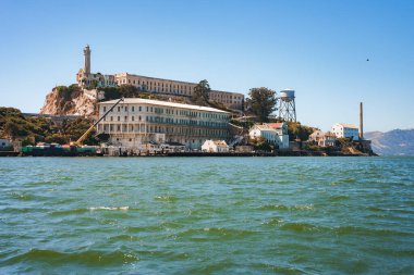 San Francisco Körfezi 'ndeki Alcatraz Adası' nın berrak, güneşli manzarası. Sudan bir fotoğraf, eski hapishane kompleksi ve ikonik deniz fenerini gösteriyor. Arkaplanda kayalar, binalar ve mavi gökyüzü.