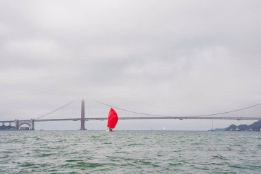 Serene view of San Francisco Bay with the Golden Gate Bridge covered by mist. A vibrant red sail yacht contrasts the calming water and muted sky colors. Ideal maritime scene. clipart