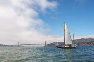 Enjoy a serene maritime view with the iconic Golden Gate Bridge in San Francisco. A yacht glides gracefully with hoisted sails on calm waters, under a clear sky. clipart