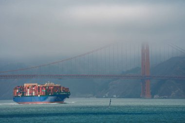 San Francisco Körfezi 'nde içi konteynırlarla dolu sisli arka planda Golden Gate Köprüsü olan, hareketli ticareti ve simgesel simgeleri temsil eden bir kargo gemisi..