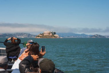 San Francisco şehir manzarasına karşı Alcatraz Adası 'nın teknesindeki turistler telefonlu fotoğraflarını çekiyor. Günlük kıyafetler, bulutlu gökyüzü, sakin okyanus manzarası. Muhtemelen tur teknesinden alınmış..