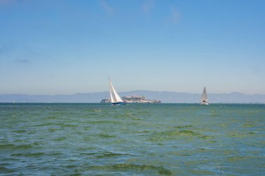 Tranquil view of San Francisco Bay with choppy waters, sailboats, Alcatraz Island silhouette, and coastal mountains in the background, capturing the bays beauty and historical significance. clipart
