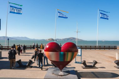 San Francisco 39 numaralı iskelede canlı bir sahne. Renkli çizgili büyük kalp heykeli aşkı simgeler. İnsanlar deniz manzarası, güneşli hava ve deniz elementlerini severler..