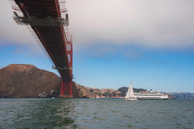 Aşağıdan Iconic Golden Gate Köprüsü, San Francisco Körfezi 'nde mavi gökyüzüne karşı yükselen kırmızı bina. Yat sakin bir şekilde süzülüyor, Marin Burnu arka planı.