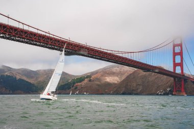 San Francisco, Kaliforniya 'daki Iconic Golden Gate Köprüsü' nün kendine özgü turuncu rengi sisle kaplıydı. Yelkenli aşağı süzülüyor, tepeler arka planda.