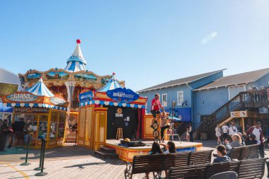 San Francisco 39 numaralı iskelede canlı bir sahne. Güneşli hava, ahşap banklar, klasik atlıkarınca, içecek standı, sokak performansı, deniz mimarisi.