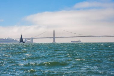 Arka planda ikonik Golden Gate Köprüsü olan San Francisco Körfezi 'nin huzurlu manzarası. Sis, yelkenli, kargo gemisi ve sakin sular güzel bir manzara yaratır..
