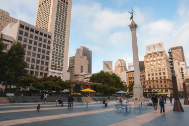 San Francisco şehir merkezindeki şehir meydanında yüksek binalar ve bir anıt var. Yayalar, açık hava koltukları ve bulutlu bir gökyüzünün altında rahat bir atmosfer..