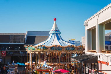 Süslü süslemeleri olan canlı klasik atlıkarınca, biniciler için atlar, mavi ve beyaz gölgelik, kırmızı tepe, ticari binalarla çevrili, muhtemelen Pier 39, San Francisco.