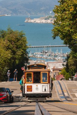 Iconic San Francisco cable car climbing a steep hill with clear tracks. Wooden car 4 offers scenic ride with views of waterfront, marina, Alcatraz Island. Sunny day, tourists waiting. clipart